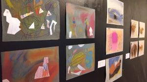 Oppilaiden pastellitöitä ripustettuna mustalle seinälle